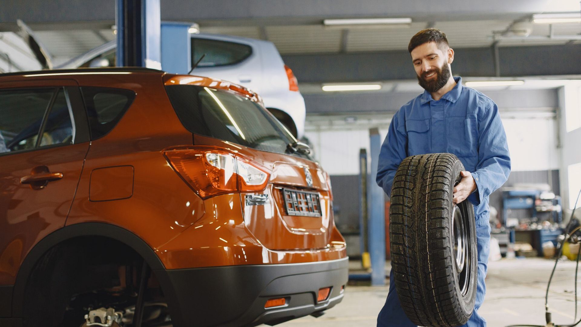 Mechaniker in blauem Overall trägt einen Reifen zu einem aufgebockten Wagen